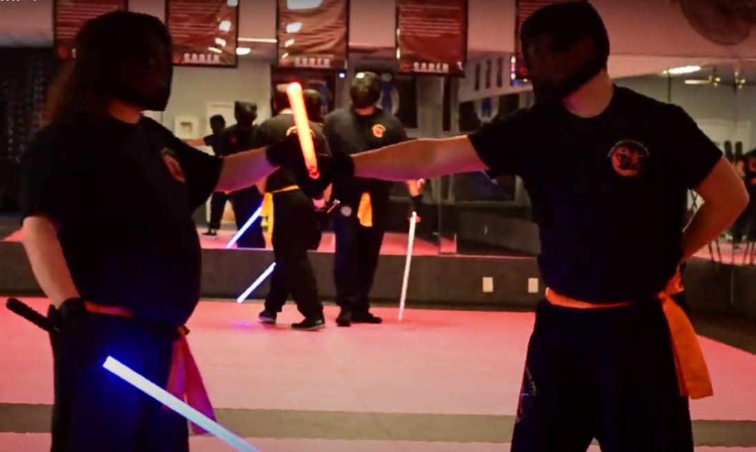 Two adult LSTA students in headgear, sabers crossed at the guard, on the academy mat