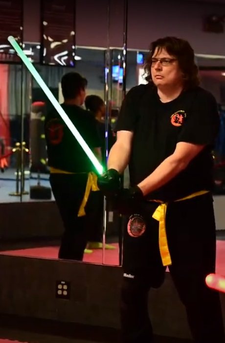 An adult LSTA student holding a lit saber in a two-handed guard beside the mirror